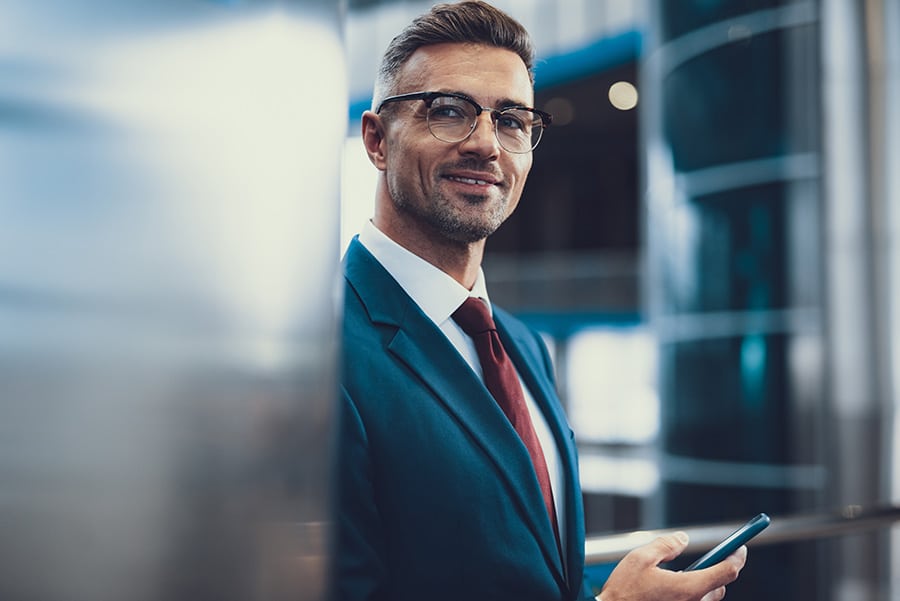 Photo of elegant adult man standing inside business center. He looking at camera and holding cellular phone in hands